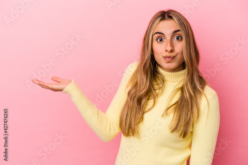 Young blonde caucasian woman isolated on pink background showing a copy space on a palm and holding another hand on waist.