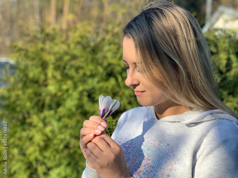 A beautiful blonde girl holding white crocus flowers in her hand, smiling and sniffing at them against the background of green tree branches. A beautiful blonde girl holding white crocus flowers in her hand, smiling and sniffing at them against the background of green tree branches.