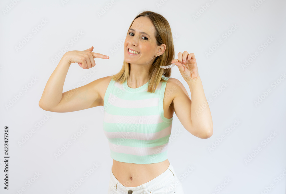 Young beautiful woman holding an invisible aligner braces against white studio background smiling and showing her perfect white teeth. Dental healthcare concept. Young beautiful woman holding an invisible aligner braces against white studio background smiling and showing her perfect white teeth. Dental healthcare concept.