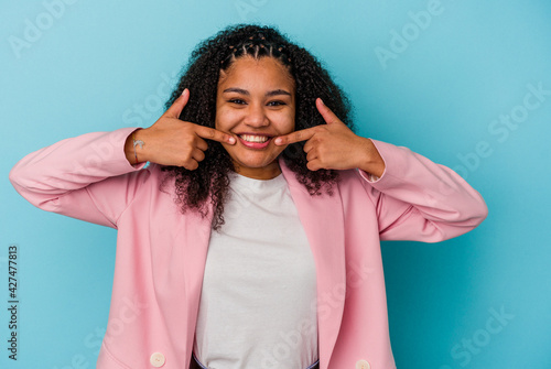 Young african american woman isolated on blue background smiles, pointing fingers at mouth.