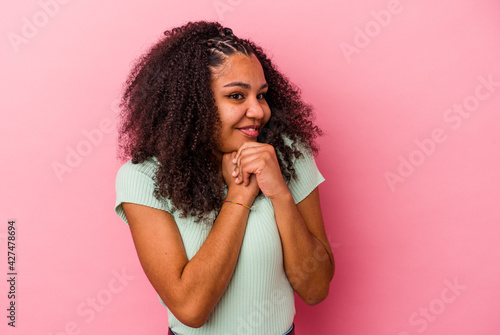 Young african american woman isolated on pink background keeps hands under chin, is looking happily aside.