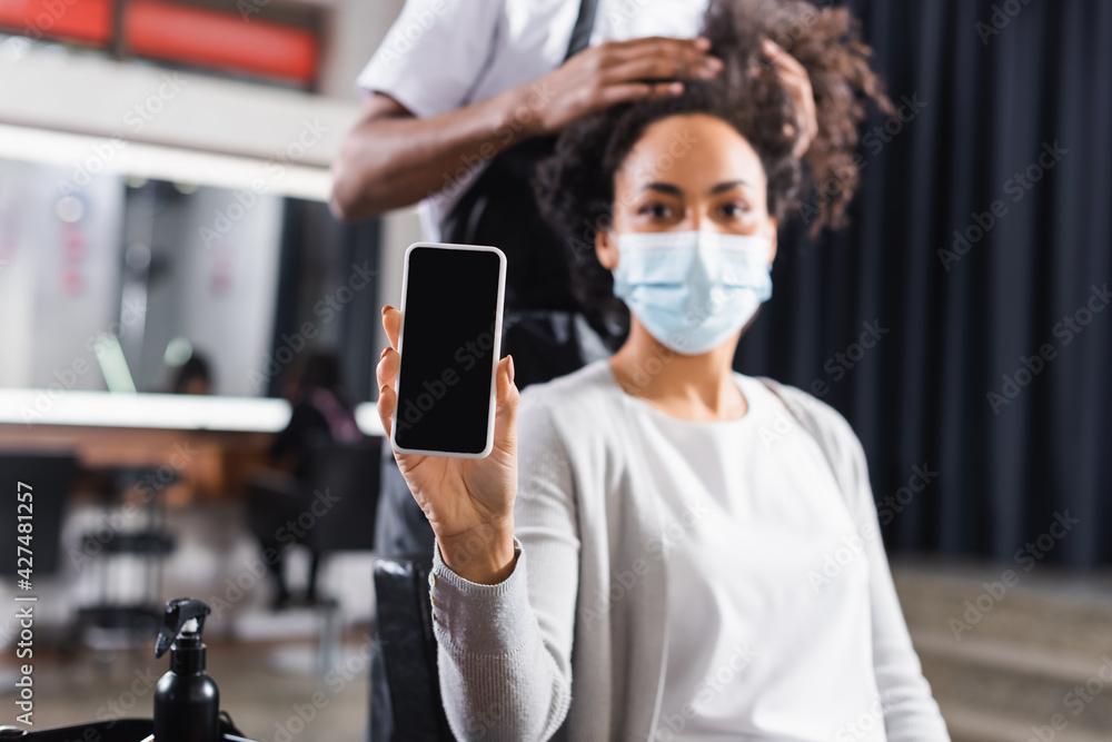 Smartphone with blank screen in hand of blurred african american client in medical mask near hairdresser