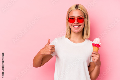 Young venezuelan woman eating an ice cream isolated on pink background smiling and raising thumb up