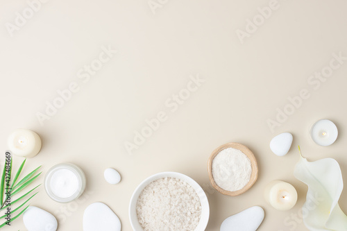 Spa background with bowl of salt, skin care products, white stones, candles, palm leaf and white flower on pastel beige. Flat lay, copy space