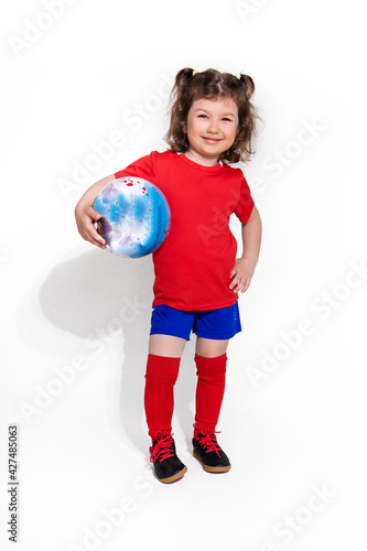 little beautiful girl in football uniform and with ball in her arm on white background looks into camera. Women's football concept for kids
