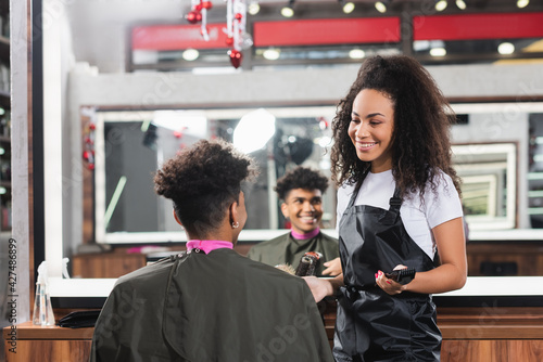 African american client sitting near smiling hairstylist with trimmer in salon
