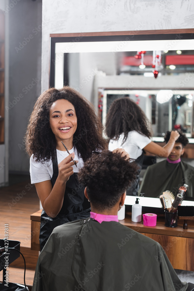 Positive african american hairdresser with comb and scissors working with client in salon - obrazy, fototapety, plakaty 