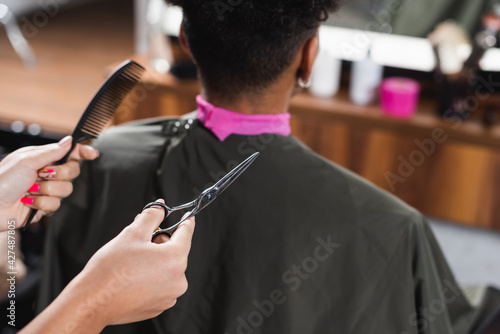 Scissors and comb in hands of hairdresser near african american man on blurred background