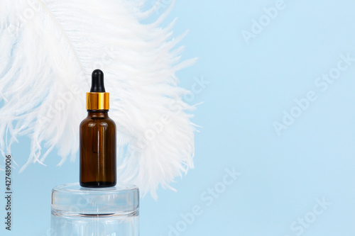 Cosmetology product for facial care. Serum in glass bottle with pipette on glass podium and large white ostrich feather on blue background. Essential oil for care of skin. Skin care, moisturizer. photo