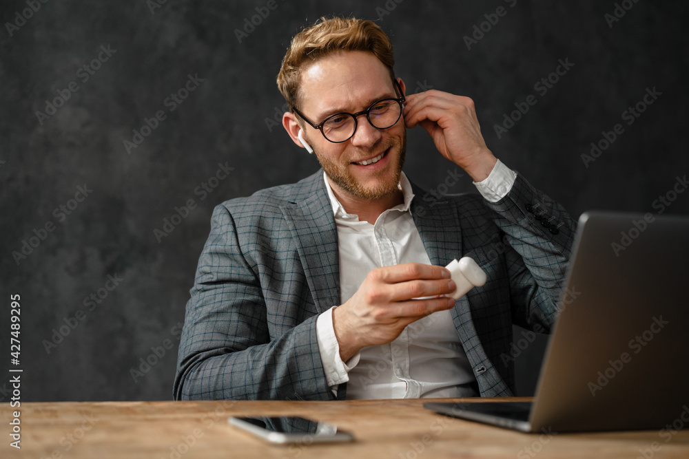 The smiling man in jacket and glasses inserting headphones into the ear in an office The smiling man in jacket and glasses inserting headphones into the ear in an office