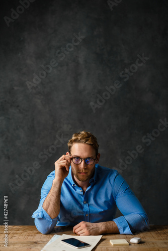 The vertical portrait of a pensive man sitting at the table and adjusting glasses in the studio
