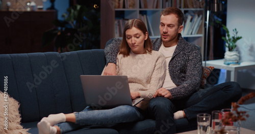 Cheerful affectionate girlfriend with boyfriend using laptop together on sofa sitting and enjoying peaceful evening. Entertainment home. Relationships.