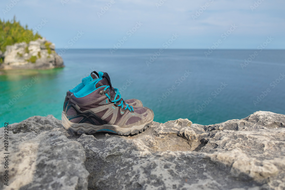 Close up of pair of old worn out weathered hiking boots on a rocky cliff over turquoise water of Georgian Bay, sunny day, selective focus, space for copy. Hiking, camping, active lifestyle concept. Close up of pair of old worn out weathered hiking boots on a rocky cliff over turquoise water of Georgian Bay, sunny day, selective focus, space for copy. Hiking, camping, active lifestyle concept.