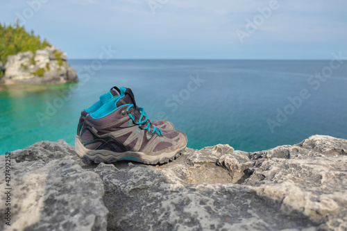 Close up of pair of old worn out weathered hiking boots on a rocky cliff over turquoise water of Georgian Bay, sunny day, selective focus, space for copy. Hiking, camping, active lifestyle concept.