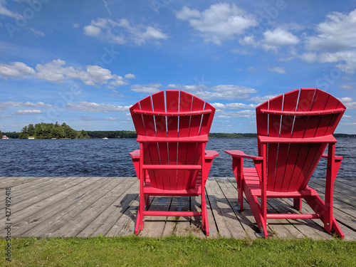 Papier Peint photo Chaises Muskoka Adirondack en bois rouge vif sur un quai  en face du lac, belle journée ensoleillée, ciel bleu avec des nuages  blancs. Muskoka, Ontario, Canada. - Nikkel-Art.fr