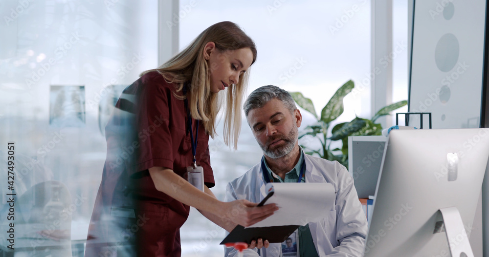 Obraz premium Young Caucasian Nurse Woman Asking Advice from Adult Doctor Negotiating in Private Medical Office. Staff Cooperation. Mixed Ethnicity. Hospitals.