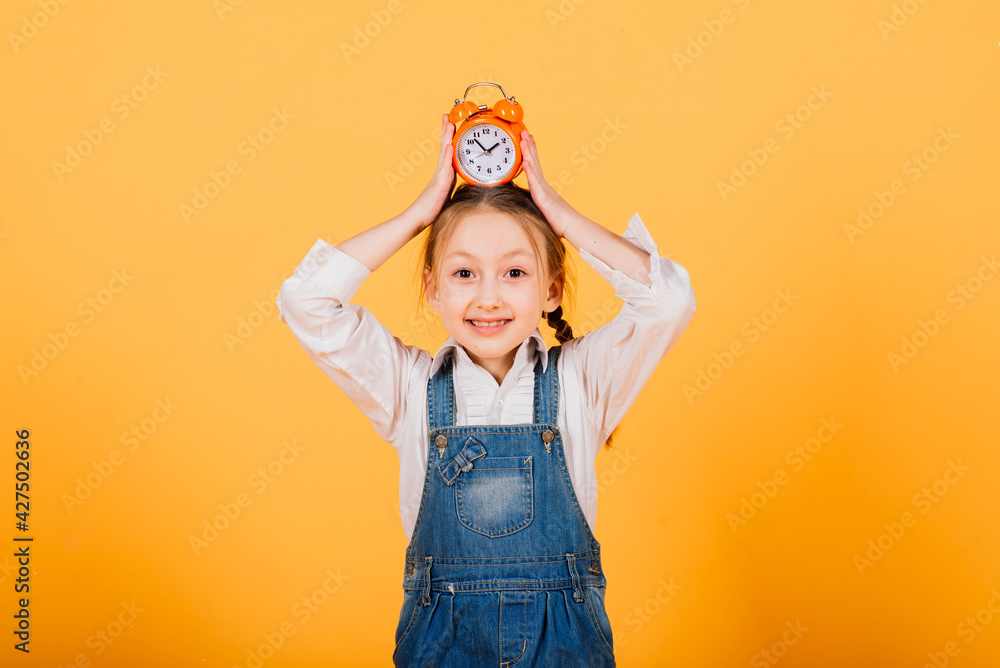 Isolated on yellow, attractive caucasian child in blue jeans with long bright hair hold alarm clock Isolated on yellow, attractive caucasian child in blue jeans with long bright hair hold alarm clock