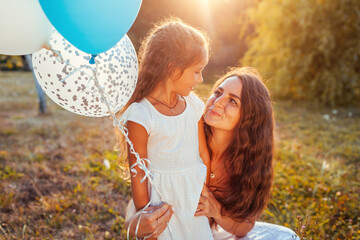 Mother's day. Little girl walking with mother and holding baloons. Family having fun in summer park. Woman hugging kid