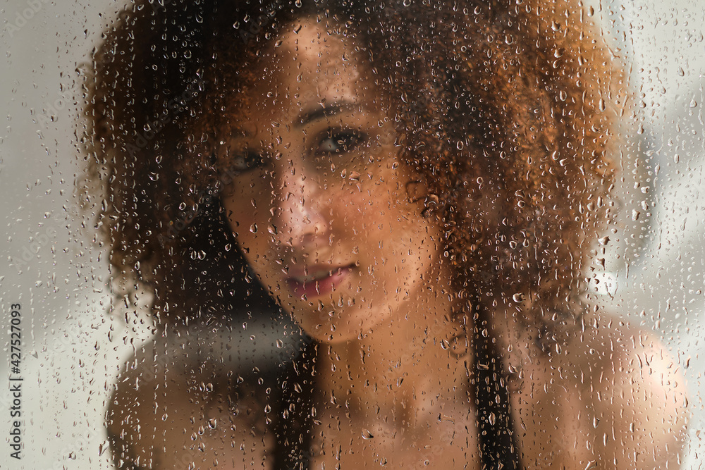 Sensual portrait of young woman taking a shower. Defocused female looks through the glass of the shower stall. Art portrait. Feminine beauty. Skincare wellness.