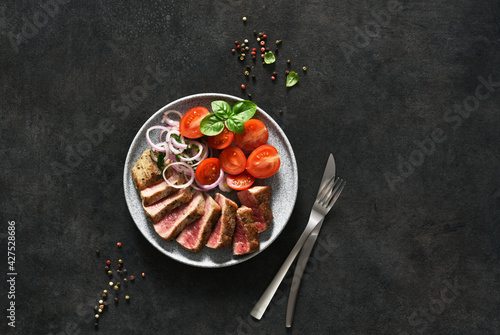 Grilled beef slices and tomatoes and onions in a concrete plate on a black background.