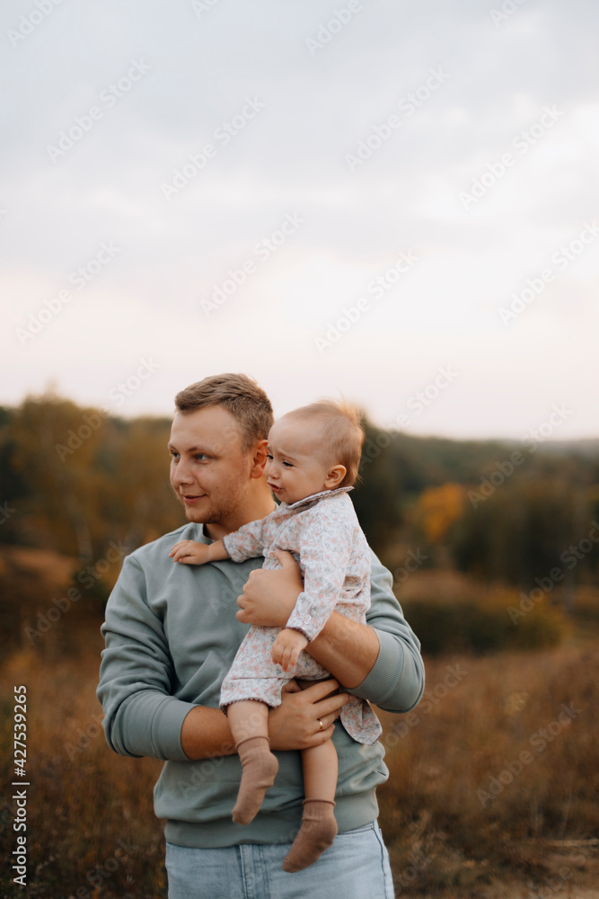 Fototapeta premium disgruntled child in dad's arms. young father with a little daughter or son in his arms. the child is about to cry in the guy's arms. tired child in his father's arms while walking in the autumn park.