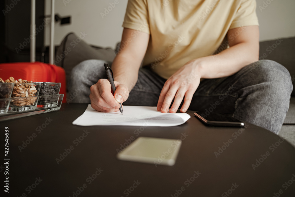 Young man traveler is sitting on couch and filling documents in hotel room on summer vacation. Cool male in casual clothing