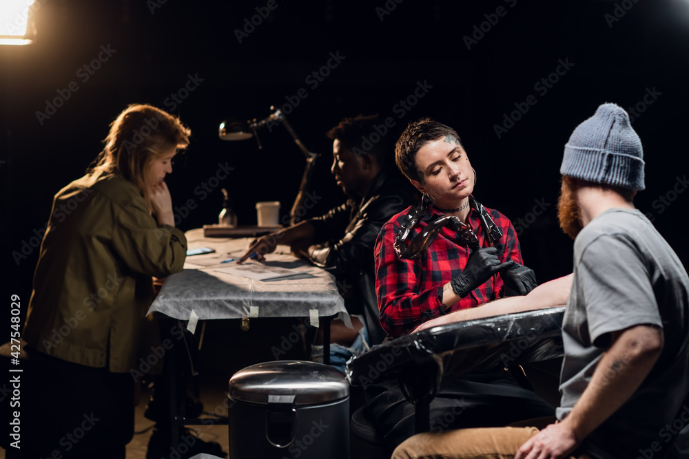 An experienced tattoo artist gives a tattoo to a white scandinavian hipster man with a red beard in his tattoo parlor. An experienced tattoo artist gives a tattoo to a white scandinavian hipster man with a red beard in his tattoo parlor.