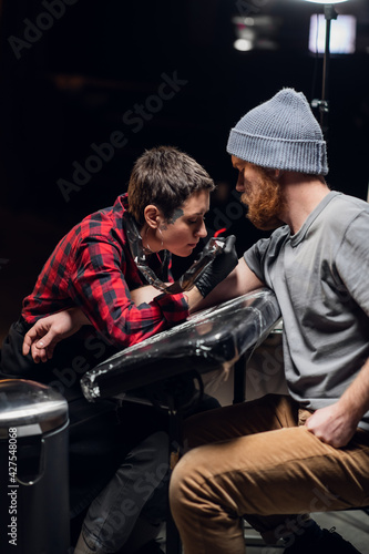 An experienced tattoo artist gives a tattoo to a white scandinavian hipster man with a red beard in his tattoo parlor.