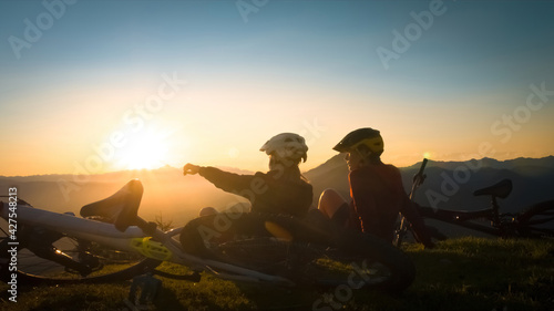 Girls sitting on mountain with bycicles looking at sunset and talking. Enjoying in beautiful nature.