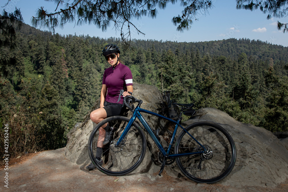 mujer latina con bicicleta descansando en el bosque, con  un paisaje de arboles al fondo. Concepto deporte al aire libre
