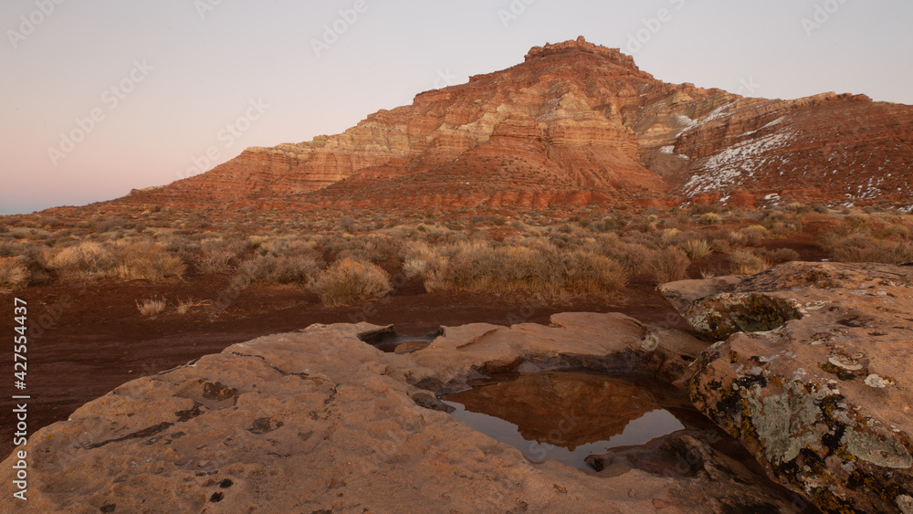 Gooseberry mesa in Southern Utah with a remnant of snow on it's northern slopes is reflected at sunset in a small puddle in a shallow slickrock depression. 