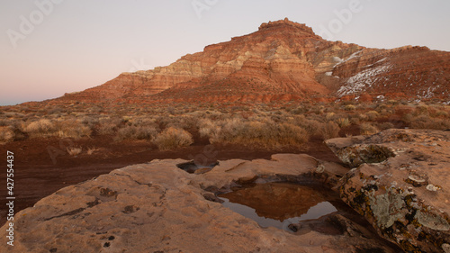 Gooseberry mesa in Southern Utah with a remnant of snow on it s northern slopes is reflected at sunset in a small puddle in a shallow slickrock depression. 