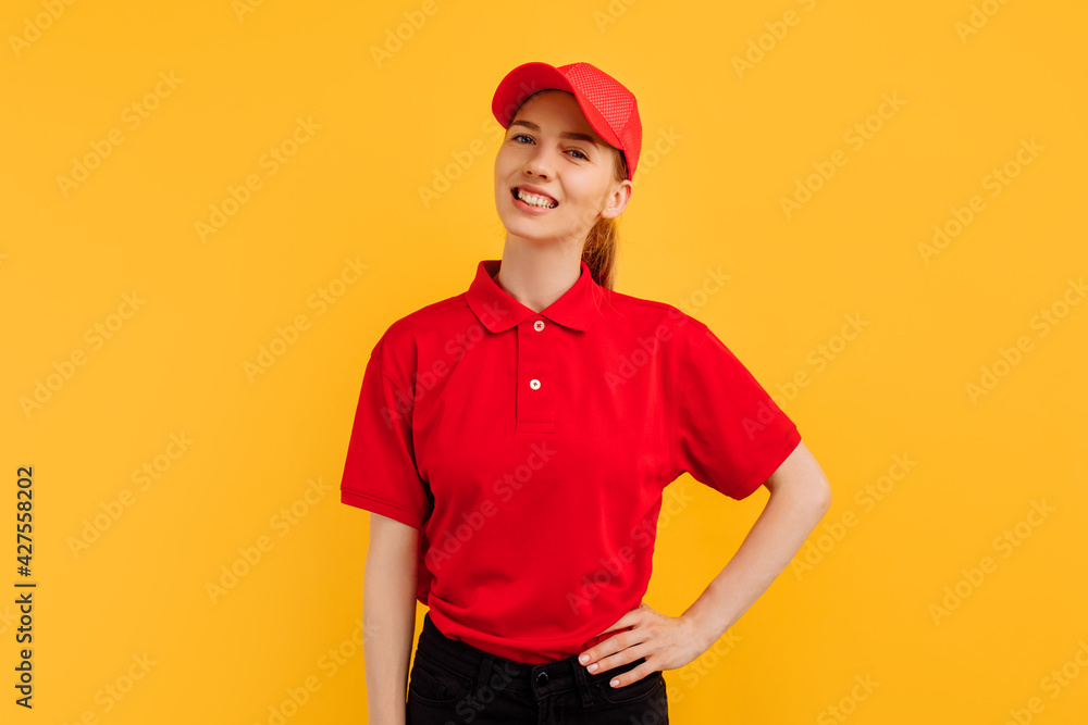 Smiling young woman courier wearing red uniform posing on yellow studio background