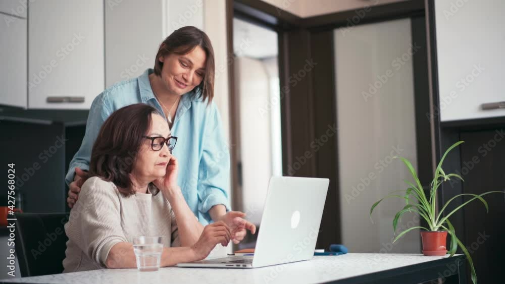 Adult daughter and her mature mother looking at the laptop screen and laughing while making online shopping. Smiling young woman teaching her parent how to use a computer. Adult daughter and her mature mother looking at the laptop screen and laughing while making online shopping. Smiling young woman teaching her parent how to use a computer.