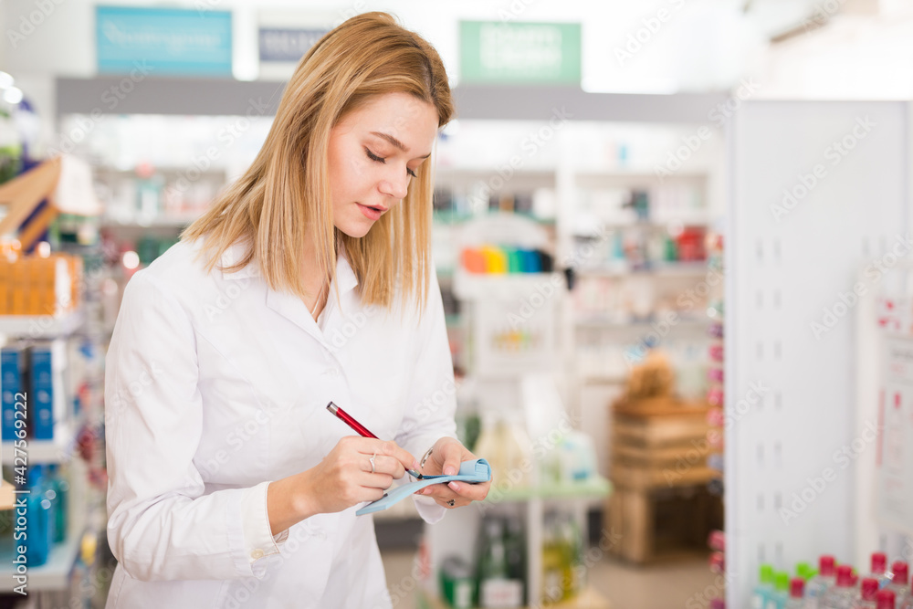 Diligent friendly smiling female pharmacist noting assortment of drugs in pharmacy