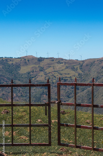 metal gate of a farm overlooking the green mountains with wind towers on top in the middle of a sunny summer day