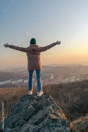 A girl in a jacket and a hat stands on a rock and looks at the city from a height during sunset