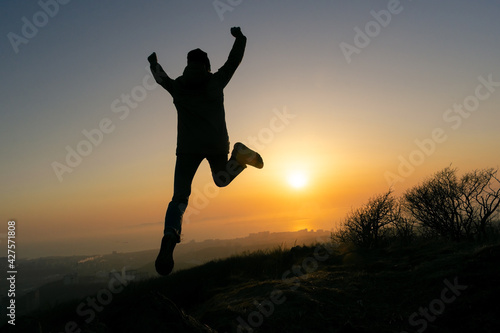 Girl jumping in a jacket and a hat on the top of the mountain during sunset. Silhouette in the rays of the sun