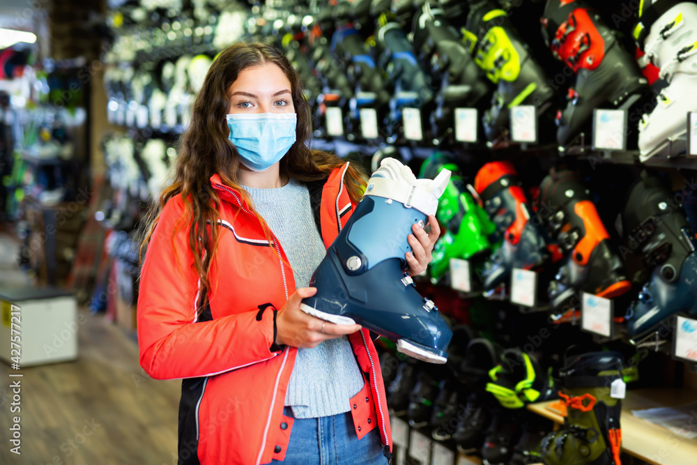Young customer woman in protective mask is choosing boots for skiing in store