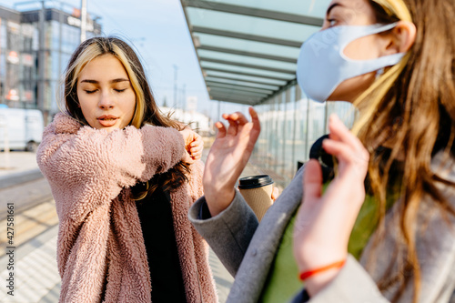 Young sick student teenager woman outside at bus stop is sneezing into the elbow by an allergy or cold. Scared woman in protective mask afraid cough woman outdoor. Keep distance. Stop corona virus