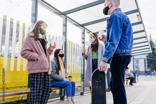 Four people waiting on bus stop for a public transport. Group of friends millennial man and women waiting drink coffee to go and talking at bus stop commuting