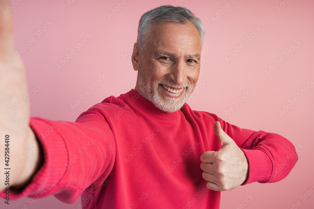 Attractive, elderly man taking a selfie, thumb up, everything is cool. Man on a pink background, copy space, place for text. Attractive, elderly man taking a selfie, thumb up, everything is cool. Man on a pink background, copy space, place for text.