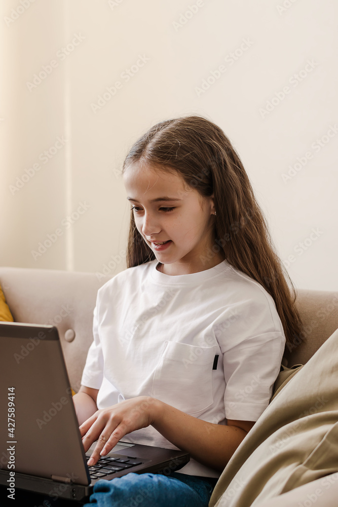 A smiling dark-haired teenage girl sitting at home on the couch,using a laptop,surfing social networks media.Distance education,leisure time. A smiling dark-haired teenage girl sitting at home on the couch,using a laptop,surfing social networks media.Distance education,leisure time.