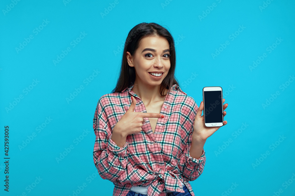 Pleased girl indicates at blank mobile screen, advertises gadget for buying, stands over blue wall Pleased girl indicates at blank mobile screen, advertises gadget for buying, stands over blue wall