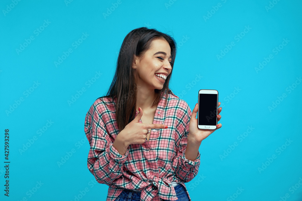Pleased girl indicates at blank mobile screen, advertises gadget for buying, stands over blue wall Pleased girl indicates at blank mobile screen, advertises gadget for buying, stands over blue wall