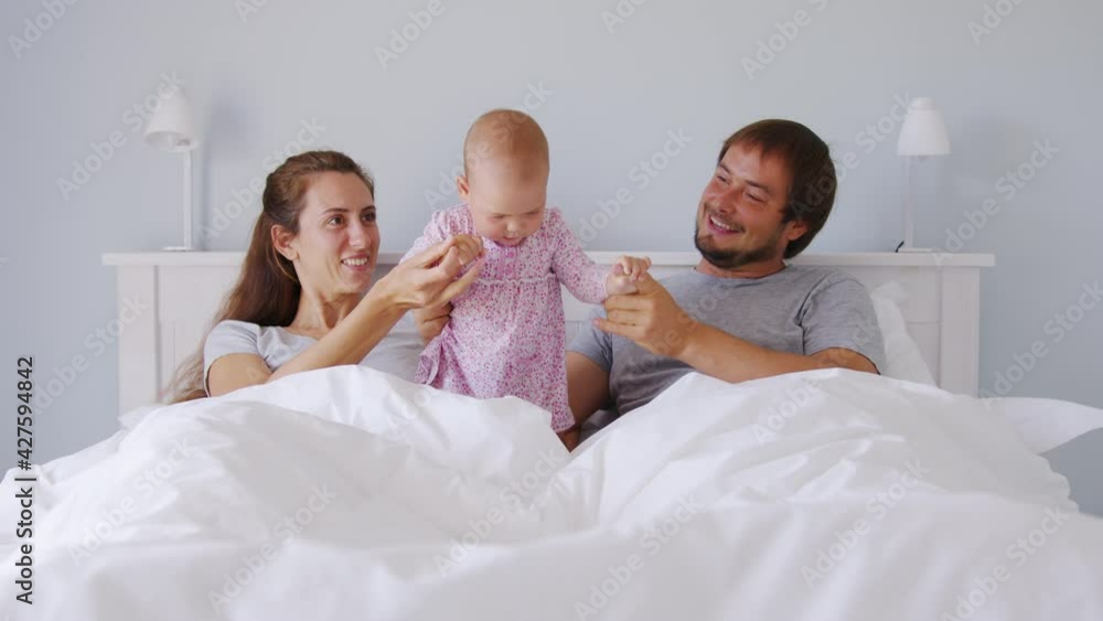 Young Family, Father, Mother and Infant Baby Together Laughing and Hugging, Lying on a Bed. White Home Interior.