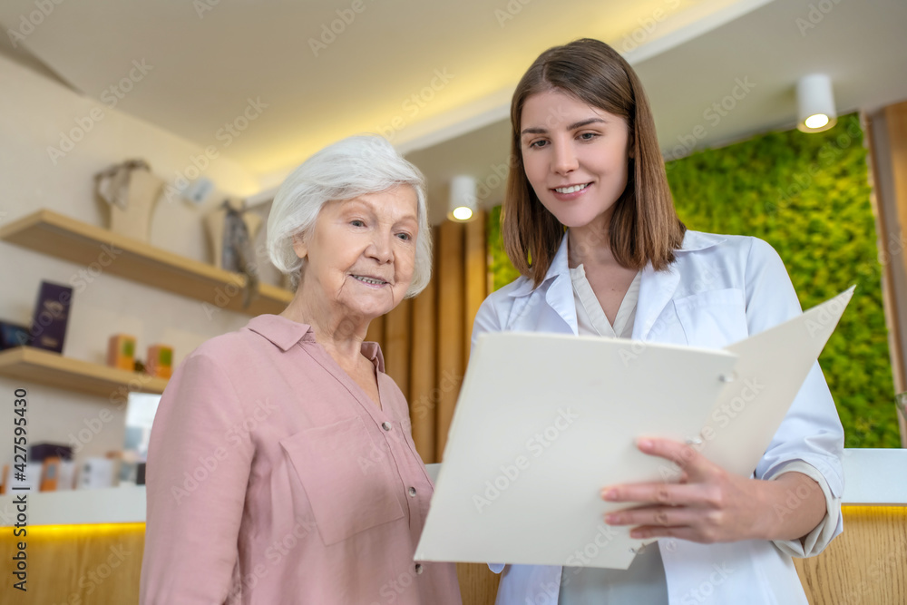 Young dark-haired cosmetologist showing the list of procedures to a customer Young dark-haired cosmetologist showing the list of procedures to a customer