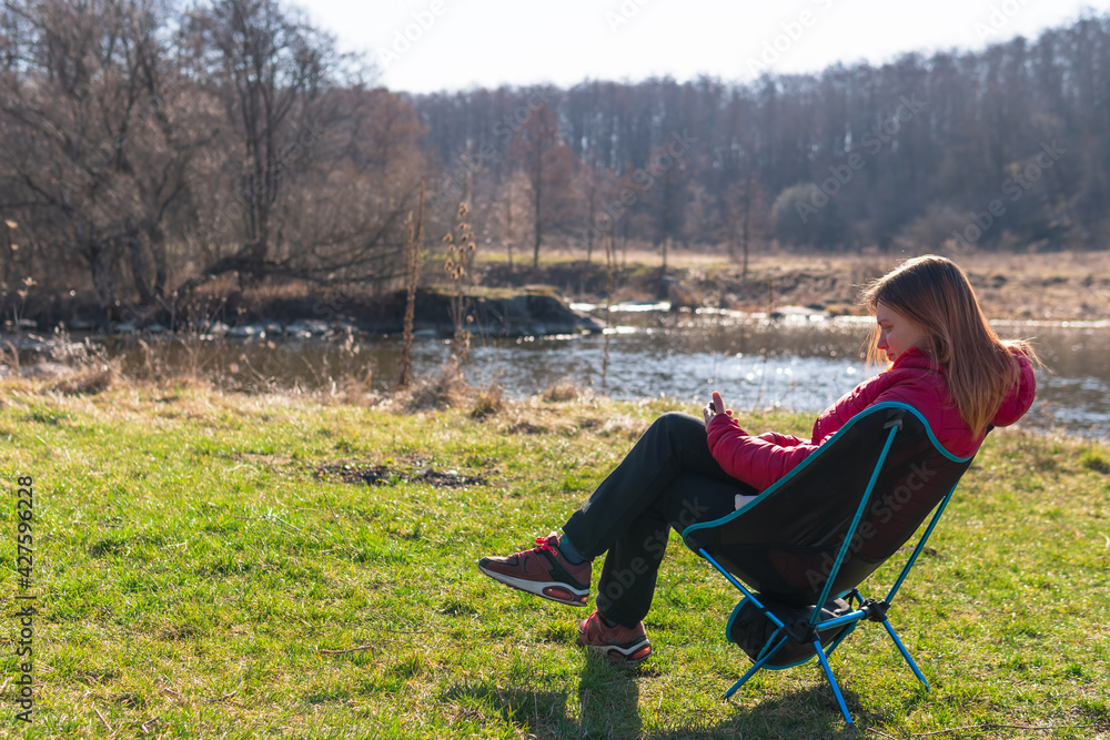 outdoor portrait of woman sitting in folding chair with smartphone near the river and forest. day time. Travel equipment. Copy space outdoor portrait of woman sitting in folding chair with smartphone near the river and forest. day time. Travel equipment. Copy space