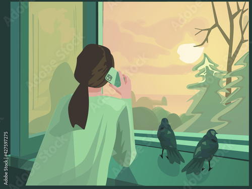 The girl stands at the window. Talking on the phone. Looks at the sunset.
