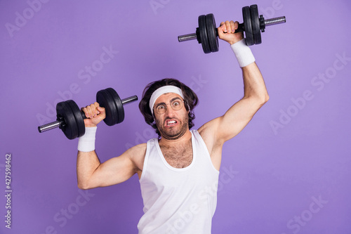 Photo of young funky funny crazy man in glasses lifting heavy dumbbell building muscles isolated on violet color background photo